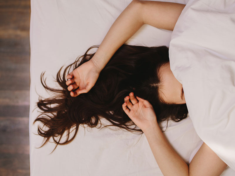 Woman's in bed with face under sheet, her hair and arms visible