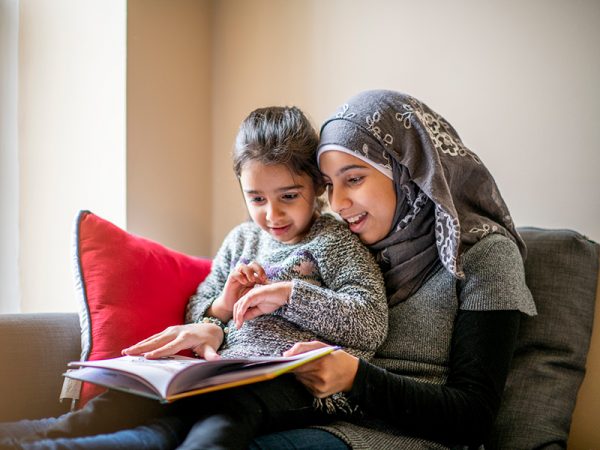 girl wearing a hijab sits on a couch with her little sister on her lap and reads her a bedtime story